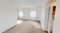 This single-floor residence offers a clean, functional layout designed for comfortable everyday living. The generous living room serves as the heart of the home, featuring plush wall-to-wall carpeting, crisp white walls, and a ceiling fan overhead. Two large windows dressed with vertical blinds flood the space with natural light, creating a bright and welcoming atmosphere. A tiled entryway provides a practical transition point, with an adjacent dining or secondary living area that opens conveniently to the kitchen.

The galley-style kitchen is well-appointed with warm honey-toned wood cabinetry lining both walls, offering ample storage and counter space. White appliances, including a gas range and refrigerator, complement the light tile flooring and bright countertops. A window above the sink adds a touch of natural light to the workspace.

The bedrooms are carpeted and feature white walls with ample closet space, including a sliding double-door closet in one room. Each room benefits from natural light through vertical-blind-covered windows. The bathrooms are finished with light tile flooring; one includes a single-sink vanity with wood cabinetry and a lighted mirror, while the other features a tub and shower combination with a sliding glass enclosure.