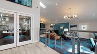 This well-appointed community clubhouse combines contemporary design with a vibrant, welcoming atmosphere. The main lounge serves as the heart of the space, featuring soaring vaulted ceilings, a striking teal geometric accent wall, and a bold mix of patterned teal and charcoal carpet tiles. A tufted caramel leather sofa, modern artwork, and a sculptural multi-globe chandelier add character and warmth. Built-in workstations along one wall provide a functional business center area within the lounge.

A split-level layout connects the main lounge to additional areas via a sleek cable-railed staircase. The entry zone features generous ceiling height, bold abstract wall art, and abundant natural light. Adjacent to the lounge is a stylish secondary lounge room with a dramatic floor-to-ceiling fireplace surround in dark tile, open display shelving, teal accent walls, light wood flooring, and a wet bar with granite countertops — ideal for social gatherings.

The expansive fitness center is outfitted with treadmills, ellipticals, stationary bikes, a full dumbbell rack, resistance machines, stability balls, and wall-length mirrors. Large windows allow natural light to fill the space, and blue-toned carpet tiles carry the design palette throughout. Together, these amenities create a cohesive, upscale communal environment.