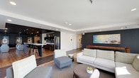 This thoughtfully designed single-floor space offers a seamless blend of social and functional areas, unified by a modern aesthetic and carefully chosen finishes. The layout flows between several distinct zones, each with its own character while maintaining visual cohesion throughout.

The lounge area anchors the space with a generous sectional sofa, accent chairs, and a round coffee table arranged on a neutral area rug. A bold dark accent wall provides a dramatic backdrop, complemented by curated artwork. Large windows on multiple sides flood the room with natural light and offer views of surrounding greenery, creating a bright and welcoming atmosphere. A wall-mounted television enhances the space's functionality.

The kitchen features a striking dual-island layout with white quartz countertops, dark tile cladding, and a row of modern black bar stools. Wood-toned cabinetry, open display shelving, and stainless steel appliances contribute to the space's polished, contemporary feel. A bank of windows above the counter frames outdoor views beautifully.

The dining area, finished in deep teal tones with pendant lighting overhead, accommodates multiple wooden tables and chairs, making it well-suited for group gatherings. Artwork adorns the walls, adding personality to this intimate setting. Hardwood-style flooring runs throughout, tying the zones together cohesively.