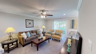 Located at Fairfield Courtyard at Lake Grove in Lake Grove, NY, this single-floor residence combines comfortable design with thoughtful finishes across a well-organized layout. The expansive living room serves as the heart of the home, featuring plush carpeting, crown molding, recessed lighting, and a ceiling fan with light fixture. A large sliding glass door fills the space with natural light and provides access to an outdoor balcony, offering pleasant views of the surrounding greenery. The kitchen is outfitted with white shaker-style cabinetry, marble-look quartz countertops, a subway tile backsplash, stainless steel appliances including a gas range and built-in microwave, and light hardwood-style flooring. A stacked washer and dryer unit is conveniently located just off the kitchen. The primary bedroom is generously sized with carpeting, crown molding, a ceiling fan, and a walk-in closet. A second bedroom features a large window with outdoor views and ample storage. The bathroom is elegantly finished with basket-weave tile flooring, a marble-top vanity, and a glass-enclosed shower-tub combination with crisp white tile surround and a natural light window above.