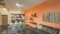 This single-floor space makes a strong visual impression through its open-concept layout, where the living, dining, and kitchen areas flow together without division. The most striking architectural feature is the bold orange accent wall in the living area, adorned with three floating display shelves that showcase decorative objects and greenery, adding depth and personality to the space.

The living area is anchored by a charcoal-gray sofa dressed with green and striped accent pillows, complemented by contemporary wall art. Polished concrete floors run throughout, lending an urban, industrial character to the space. Track lighting and pendant fixtures provide layered illumination across the open floor plan.

The dining area features a glass-top table paired with transparent acrylic chairs, contributing to a modern aesthetic while keeping the space visually open. Adjacent to the dining space, the kitchen is outfitted with stainless steel appliances, including a refrigerator, range, and microwave, set against dark cabinetry. Pendant lights hang above, tying the kitchen visually into the broader living space. Overall, the space balances bold color choices with contemporary furnishings to create a cohesive and energetic environment.