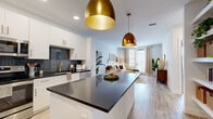 This well-appointed 1-bedroom, 1-bathroom unit offers a cohesive open-concept layout that maximizes its efficient footprint. Light wood-look flooring flows throughout, tying together each area seamlessly. The kitchen is a standout feature, showcasing crisp white cabinetry with gold hardware, a bold dark blue tile backsplash, dark quartz countertops, stainless steel appliances, and a central island illuminated by striking brass pendant lights. Built-in open shelving along one wall adds both function and visual interest. The kitchen opens directly into the combined dining and living area, where natural light pours in through large windows and a glass door. The living space is anchored by a ceiling fan and offers a comfortable, airy ambiance. The bedroom features a calming gray accent wall, carpeted floors, and windows that invite ample natural light. A dedicated closet provides organized storage. The bathroom is finished with a large framed mirror, dark quartz vanity countertop, white cabinetry with gold hardware, and floor-to-ceiling gray tile in the shower surround. The unit's consistent use of white walls, warm wood tones, and gold accents creates a modern yet inviting atmosphere throughout.