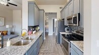 This single-floor residence offers a thoughtfully designed layout that balances style and functionality across its key living spaces. The galley-style kitchen is a standout feature, showcasing slate-blue shaker cabinetry, granite countertops, a double stainless steel sink, a mosaic tile backsplash, and a full suite of stainless steel appliances including a refrigerator, electric range, and over-the-range microwave. Wood-look plank flooring runs throughout, providing a cohesive and modern aesthetic.

The living room is bright and welcoming, featuring elevated ceilings, a ceiling fan, and large windows that allow natural light to fill the space. The neutral warm-gray color palette creates a calm and inviting ambiance throughout.

The space includes two bedrooms, each equipped with a ceiling fan and windows offering natural light. One bedroom features a more spacious layout with a dresser alcove, while the other offers a cozy, intimate feel with a tufted upholstered headboard as a focal point.

The full bathroom is elegantly finished with a granite-topped blue vanity, wood-look flooring, and an arched-niche tub and shower combination with tile surround. A dedicated in-unit laundry closet with side-by-side washer and dryer and built-in shelving adds everyday convenience to this well-rounded residence.