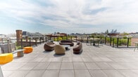 Located at 1700–1706 N Howard St in Philadelphia, PA, this contemporary rooftop amenity space seamlessly blends indoor comfort with outdoor living. The building's exterior is clad in corrugated metal panels, giving it a distinctly modern, industrial aesthetic that complements the surrounding neighborhood.

The interior living area is generously proportioned, featuring large floor-to-ceiling windows and a striking retractable glass garage-style ceiling panel that floods the space with natural light and opens the room directly to the terrace. A textured wood-plank accent wall serves as a bold focal point, complemented by large-format tile flooring throughout. Seating areas are arranged to encourage social gathering, with vibrant modular sectionals in bold orange and magenta tones, neutral sectional seating with yellow accent pillows, curved lounge chairs in olive green and orange, and a marble-topped counter adding a refined touch. Curated artwork and architectural lighting fixtures complete the interior's energetic, design-forward character.

The expansive outdoor terrace wraps the building and offers multiple zones for relaxation and dining, including sculptural lounge furniture, dining tables with seating, and chaise lounges. Perimeter cable railings ensure unobstructed panoramic views of the Philadelphia skyline and surrounding neighborhoods, making this an exceptional rooftop destination.