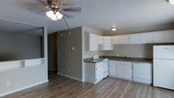 This single-floor space offers a practical and straightforward layout designed for comfortable everyday living. The open-concept design connects the main living area to the kitchen, creating a cohesive flow throughout the unit. Hardwood-style flooring runs continuously across the space, adding warmth and visual continuity to the neutral color palette of taupe and white tones.

The kitchen is positioned along one wall and features white cabinetry with ample upper and lower storage, dark countertops, a white electric range, and a double-basin sink beneath a flush-mounted ceiling light. The cabinetry layout follows an L-shaped configuration, maximizing the use of available counter and storage space.

The living area is equipped with a ceiling fan and light fixture, providing both air circulation and overhead illumination. A textured popcorn ceiling is present throughout, and the walls are painted in a warm greige tone that complements the flooring. A hallway visible in the background suggests additional rooms beyond the main living and kitchen space. The overall ambiance is clean, functional, and move-in ready.