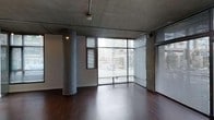 This single-floor space embodies the character of contemporary urban industrial design. The exposed concrete ceiling sets a raw, unfinished aesthetic that is immediately striking upon entry, complemented by a bold structural column finished in matching concrete — a hallmark of loft-style architecture. Dark hardwood flooring grounds the space with warmth, creating a compelling contrast against the cool tones of the concrete overhead.

Natural light pours in abundantly through expansive floor-to-ceiling corner windows, which wrap around two sides of the space to offer dynamic, unobstructed views of the surrounding cityscape. The large glazed panels are fitted with horizontal metal blinds, allowing for flexible light control while maintaining the industrial character of the interior. The open, column-free layout — aside from the singular central pillar — provides a versatile and spacious footprint adaptable to a range of uses.

Exposed ceiling-mounted lighting fixtures and visible utility infrastructure further reinforce the industrial aesthetic. The overall ambiance is modern and urban, with a clean, open feel that makes the space feel bright and expansive despite its single-story configuration. This is a well-defined space that balances raw architectural detail with practical functionality.