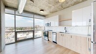 Located at 240–246 S Vandeventer Ave in St. Louis, MO, this 1-bedroom, 1-bathroom unit showcases a striking industrial-modern aesthetic with exposed concrete ceilings, visible ductwork, and light hardwood flooring throughout. The open-concept living, dining, and kitchen area is anchored by dramatic floor-to-ceiling windows that bathe the space in natural light and frame expansive views of the St. Louis skyline and surrounding neighborhoods. The kitchen is thoughtfully appointed with light wood cabinetry, gloss white upper cabinets, a marble-style backsplash, quartz countertops, stainless steel appliances including a refrigerator, range, microwave, and dishwasher, and a matte black faucet at the undermount sink. An open shelf adds functional display space. The living area is generously proportioned with panoramic city views as a focal point. The bathroom features matching light wood vanity cabinetry, a white countertop, matte black hardware, tile flooring, and an adjacent laundry closet with built-in shelving. The bedroom offers a comfortable, private retreat. Throughout, the interplay of raw concrete, warm wood tones, and clean white finishes creates a refined urban living environment.