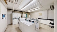 Located at 570 W Dekalb Pike in King of Prussia, PA, this shared laundry facility is a well-equipped, single-level space designed to efficiently serve its users. The room is outfitted with multiple top-loading and front-loading washing machines, as well as stacked dryer units, providing ample capacity for residents. Bright fluorescent lighting runs along the ceiling, illuminating the white-tiled floor and light-colored walls to create a clean, functional atmosphere. Overhead, exposed pipes and utility lines reflect the room's utilitarian design. A utility sink is available for hand-washing or pre-treating garments, accompanied by a dark-countered folding station ideal for sorting and organizing laundry. A small seating area with metal chairs offers a comfortable place to wait during wash cycles. Informational notices and digital displays are mounted on the walls, keeping users informed about machine operation and facility guidelines. A waste bin is conveniently placed within the space. Decorative artwork on the walls adds a subtle touch of character to the otherwise practical environment. Overall, this laundry facility is thoughtfully arranged to maximize usability and comfort for all who utilize it.