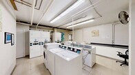 Located at 570 W Dekalb Pike in King of Prussia, PA, this shared laundry facility is a well-equipped, single-level space designed to efficiently serve its users. The room is outfitted with multiple top-loading and front-loading washing machines, as well as stacked dryer units, providing ample capacity for residents. Bright fluorescent lighting runs along the ceiling, illuminating the white-tiled floor and light-colored walls to create a clean, functional atmosphere. Overhead, exposed pipes and utility lines reflect the room's utilitarian design. A utility sink is available for hand-washing or pre-treating garments, accompanied by a dark-countered folding station ideal for sorting and organizing laundry. A small seating area with metal chairs offers a comfortable place to wait during wash cycles. Informational notices and digital displays are mounted on the walls, keeping users informed about machine operation and facility guidelines. A waste bin is conveniently placed within the space. Decorative artwork on the walls adds a subtle touch of character to the otherwise practical environment. Overall, this laundry facility is thoughtfully arranged to maximize usability and comfort for all who utilize it.