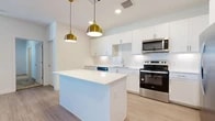 This thoughtfully designed single-floor home features 2 bedrooms, 2 bathrooms, and two closets, delivering a clean, modern aesthetic throughout. The open-concept kitchen and living area serves as the heart of the home, showcasing crisp white shaker cabinetry, a textured tile backsplash, quartz countertops, and a central island illuminated by striking gold pendant lights. A full suite of stainless steel appliances — including a range, microwave, dishwasher, and French-door refrigerator — completes the kitchen. Light wood-tone flooring runs throughout the main living areas, enhancing the bright and airy atmosphere. The spacious primary bedroom includes a ceiling fan, natural light from a window with blinds, and access to a generously sized walk-in closet with built-in shelving and carpet flooring. The second bedroom also features carpet flooring and a window with outdoor views. Both bathrooms are finished with white cabinetry, quartz countertops, large vanity mirrors with overhead lighting, and wood-tone flooring. The overall design emphasizes a neutral, contemporary palette, offering a clean and functional living environment throughout.