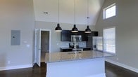 This single-story space showcases a thoughtfully designed open-concept kitchen that serves as the focal point of the layout. The kitchen is anchored by a large island topped with speckled granite countertops and an undermount sink, offering both functional workspace and a natural gathering area. Three matte black dome pendant lights hang dramatically above the island, adding a bold, modern accent against the soaring vaulted ceiling that draws the eye upward and amplifies the sense of openness.

Dark espresso cabinetry lines the back wall, providing strong visual contrast against the light gray walls and classic subway tile backsplash. Stainless steel appliances, including a range, microwave, and refrigerator, complement the contemporary aesthetic. Natural light filters in through windows positioned high on the walls, taking full advantage of the vaulted ceiling height to brighten the space.

Dark hardwood flooring runs throughout, grounding the space and adding warmth. A doorway visible to the left suggests a seamless flow into adjacent rooms, reinforcing the open and connected nature of the layout. Overall, the space balances modern design with practical functionality.