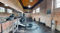This fitness center occupies a single-floor exercise room with a striking industrial aesthetic that sets it apart from a typical gym. Exposed red brick walls rise to soaring heights, culminating in a warm, wood-beam ceiling accented by pendant lights and ceiling fans that ensure both style and comfort during workouts. The polished concrete flooring adds to the raw, industrial character of the space while remaining practical and durable underfoot.

Natural light streams in through a series of large, multi-pane windows positioned along the upper walls, creating a bright and energizing atmosphere throughout the day. A full-length mirror along one wall enhances the sense of openness and supports form-checking during exercise.

The space is comprehensively equipped for a wide range of training modalities. Strength training is well supported with a dumbbell rack, adjustable benches, a cable machine, and a dedicated barbell station. Cardio options include a treadmill and a rowing machine. For functional and combat training, a freestanding heavy punching bag is positioned in its own area alongside a training mat. Kettlebells of varying weights are neatly lined along the wall for additional versatility. Roll-up garage-style doors and dark green entry doors add to the converted-warehouse charm, completing an exercise space that is both functional and visually distinctive.