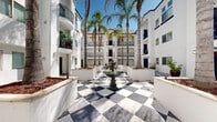 This outdoor courtyard showcases a distinctive Mediterranean-inspired architectural design, centered around a bold, large-scale black-and-white checkered stone floor that immediately commands attention. At the heart of the courtyard stands a classic stone fountain, serving as the visual focal point and lending an elegant, timeless character to the space.

Tall palm trees rise dramatically from substantial white stucco planters positioned throughout the courtyard, adding vertical dimension and a tropical ambiance. The surrounding multi-story buildings feature crisp white stucco exteriors, arched passageways, and wrought iron balconies that frame the open-air space on multiple sides, creating a cohesive and enclosed atmosphere while still allowing abundant natural light and open sky views above.

Covered walkways with arched entryways line the perimeter at ground level, providing sheltered transitions between the courtyard and interior corridors. Decorative wall-mounted lanterns and potted plants accent the pathways, adding warmth and detail to the architectural composition. The combination of geometric flooring, lush tropical greenery, and classic Southern California stucco architecture creates a vibrant, resort-like communal environment that is both visually striking and inviting.