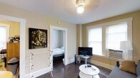 This single-floor space features a warm and functional layout with distinct living and sleeping areas. The living room is finished with rich, dark hardwood floors that add depth and character to the space. Neutral beige walls create a calm backdrop, while a ceiling-mounted light fixture provides ambient overhead lighting. A double-hung window fitted with blinds allows natural light to fill the room, enhancing its open feel. The living area accommodates a flat-screen television on a compact stand and a round white coffee table, making it a practical everyday space. A doorway with white-trimmed framing opens directly into the bedroom, which is visible from the living area and features a bed dressed in light-colored linens. The bedroom benefits from its own window, admitting additional natural light. White baseboards throughout provide a clean contrast against the dark flooring. A decorative artwork piece featuring a Ganesha motif adds a distinctive cultural touch to the living room wall. Overall, the space offers a straightforward and livable environment with a cohesive, understated aesthetic.