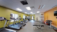 This fitness center offers a comprehensive workout environment designed to accommodate a wide variety of exercise routines. The space features a clean, modern aesthetic with bold chartreuse-yellow accent walls that energize the room, contrasted against a neutral drop-tile ceiling fitted with recessed panel lighting that ensures the area is evenly and brightly lit throughout. Durable, low-pile fitness flooring covers the ground, providing traction and cushioning ideal for a gym setting. The cardio section is well-stocked with multiple treadmills and stationary bikes, giving members ample options for aerobic exercise. A multi-station cable and weight machine anchors the strength training area, complemented by a dedicated free weight bench, offering a complete resistance training setup. Additional cable and weight machines are positioned toward the back of the room. Windows along one wall allow for natural light to supplement the overhead lighting, contributing to an open and welcoming atmosphere. A water fountain is conveniently accessible near the rear exit, and clear emergency exit signage ensures safety throughout. The single-floor layout is efficiently organized, making navigation between equipment stations intuitive and unobstructed.