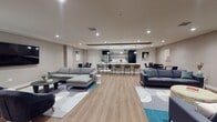 Located in the heart of Hollywood at 1717 N Cherokee Ave, this single-floor communal space is thoughtfully designed to foster social interaction and relaxed living. The expansive open-plan layout is anchored by warm wood-look flooring that flows seamlessly throughout, unifying the various functional zones under recessed lighting that provides even, ambient illumination across the entire floor.

The lounge area is generously proportioned, featuring multiple seating arrangements including plush gray sectionals, navy accent sofas, and contemporary accent chairs arranged over bold, patterned area rugs in teal, navy, and gray. A large wall-mounted flat-screen television serves as a focal point in one of the lounge zones, while curated wall art adds a refined, residential character to the space.

The centrally located kitchen is a standout feature, outfitted with matte black cabinetry, an extended white quartz island with bar seating, stainless steel appliances including a full-size refrigerator, open wood shelving, and a patterned tile floor that distinguishes the kitchen zone. Adjacent dining areas with round tables and navy chairs offer additional spaces for gathering. A distinctive cork accent wall near the entrance adds a tactile, design-forward touch to the overall aesthetic.