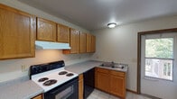 Located at 308 Rublein St in Marquette, Michigan, this single-floor unit offers a practical and straightforward layout with clearly defined living areas. The kitchen is well-equipped with warm oak cabinetry extending to the ceiling for generous storage, grey laminate countertops arranged in an L-shaped configuration, a coil-burner electric range, dishwasher, refrigerator, and a double-basin stainless steel sink. Light tile flooring runs throughout the kitchen, adding a clean, easy-to-maintain surface underfoot. The living area is spacious and carpeted, providing a comfortable, quiet atmosphere with room for a variety of furniture arrangements. Multiple carpeted rooms serve as versatile spaces, each featuring windows with blinds that invite natural light and offer views of surrounding greenery. Baseboard heating units are present in the rooms, ensuring reliable warmth during Michigan's cold winters. The bathroom is clean and functional, featuring a full tub-shower combo, a wood-framed vanity mirror, grey speckled countertop, and tile flooring that complements the rest of the unit. Throughout the space, neutral warm-toned walls and consistent trim work give the unit a cohesive, move-in-ready appearance.