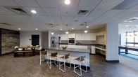 This single-floor space centers around a spacious, thoughtfully designed communal kitchen that serves as its primary feature. The kitchen is anchored by a generously sized island with a dark gray countertop, surrounded by modern white metal-framed bar stools with wood-accented seats, offering a practical and social gathering point. Matching dark countertops line the perimeter, contrasting cleanly against an abundance of white cabinetry that extends across the walls, providing substantial storage throughout. Stainless steel appliances — including a double wall oven, a large French door refrigerator, and a sink along the side wall — equip the space for serious food preparation. The drop ceiling features a grid of recessed lighting alongside pendant lights that hang above the island, ensuring the space is well-lit throughout. The flooring is a wood-toned laminate or vinyl plank that adds warmth to the otherwise neutral palette of white and gray tones. A dining area with a round table and chairs is partially visible in the background, extending the functionality of the space for group meals or meetings. The overall ambiance is clean, modern, and functional.