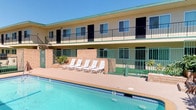 This two-story property is designed around a central courtyard that serves as the heart of the outdoor amenity space. The exterior facade features a warm stucco finish complemented by green-trimmed railings and balconies that line the upper level, providing direct access to individual units and offering views of the courtyard below. A full-length outdoor swimming pool anchors the main courtyard area, bordered by a spacious sun deck furnished with a row of white lounge chairs — ideal for relaxing under open skies. The pool is framed by decorative brick planters with tropical greenery, adding a touch of lush landscaping to the setting. A separate, octagonal spa or hot tub is tucked into a secondary enclosed courtyard area, offering a more intimate retreat. This area is accessed through green iron-gated fencing and features additional seating, along with a staircase connecting to the upper level. The property's overall aesthetic reflects a classic, sun-drenched hospitality design, with green-accented metalwork throughout providing a cohesive visual identity. Ample natural light and open-air circulation make the outdoor spaces inviting and functional year-round.