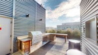 This rooftop terrace is a well-appointed outdoor amenity space set atop a modern urban building, offering striking city skyline views and an open-air atmosphere. The terrace is framed by corrugated metal cladding that lends an industrial yet contemporary character throughout. Paved in large-format stone tiles, the surface provides a clean, durable foundation for the various outdoor features.

A dedicated outdoor kitchen area serves as a central highlight, equipped with a stainless steel built-in grill set into a wood-framed counter structure, ideal for grilling and entertaining. Nearby, built-in bench seating and additional counter space offer practical areas for dining and gathering. Ornamental grasses planted in dark rectangular planter boxes add a touch of greenery and texture against the hardscape surroundings.

Passageways between the rooftop mechanical enclosures create defined corridors that open onto the main terrace, with signage indicating stairwell and elevator machine room access. Glass-paneled entry doors provide access to the rooftop and feature posted rules and regulations for resident use. Overhead, the open sky fills the space with natural light, while urban rooftops and city buildings visible on the horizon reinforce the elevated, metropolitan setting. The overall ambiance is modern, functional, and well-suited for communal outdoor use.