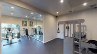 This single-story fitness center in Nederland, Texas, offers a thoughtfully designed workout environment spread across a well-organized open floor plan. The space is divided into distinct functional zones, each catering to different aspects of physical training. Recessed lighting throughout provides consistent, even illumination, complementing the neutral beige and gray tones of the walls and ceilings.

The strength training area is anchored by Quantum cable machines, offering versatile resistance training options. A comprehensive dumbbell rack lines one wall, providing a full range of free weights for varied exercise routines. Floor-to-ceiling mirrors span a significant portion of the walls, visually expanding the space while allowing users to monitor their form during workouts.

A dedicated cardio zone features a treadmill and elliptical trainer, positioned alongside large windows that offer views of the outdoor surroundings, including a playground and landscaped greenery. The natural light from these windows brightens the area and creates an inviting atmosphere.

Dark rubber flooring covers the entire space, providing durability, traction, and impact absorption suitable for high-activity use. Direct exterior access via a glass-panel door connects the fitness center to the outdoor area, enhancing convenience and ventilation. The overall design is clean, functional, and well-maintained.