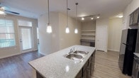 This single-story, 3-bedroom, 2-bathroom residence delivers a practical and modern living experience through a thoughtfully designed open floor plan. The kitchen serves as a central hub of the home, featuring dark espresso cabinetry that extends to ceiling height, granite-style countertops, a double-basin stainless steel sink set within a large island, and black appliances including a gas range, refrigerator, and microwave. Pendant lighting and track lighting illuminate the space, while open shelving provides additional display or storage options nearby. Wood-look plank flooring runs throughout the main living areas and bedrooms, lending a cohesive, contemporary aesthetic. Each bedroom is comfortably sized and includes ceiling fans, window blinds for natural light control, and access to ample closet space. The primary suite benefits from a spacious walk-in closet with multi-tier wire shelving. Both bathrooms are finished with granite-top dark vanities and tile flooring. The primary bath features a dual-sink vanity, while the secondary bath offers a full tub-shower combination with overhead cabinetry. A dedicated laundry area houses a full-size top-load washer with wire shelving above. Neutral wall tones and clean architectural lines throughout create a bright, move-in-ready atmosphere.