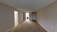 This single-floor residence offers a straightforward and functional layout designed for comfortable everyday living. The main living area is generously sized, featuring wall-to-wall carpet flooring in a warm beige tone that adds a sense of coziness to the space. Neutral gray-beige walls provide a clean, versatile backdrop throughout, complemented by crisp white baseboards and trim that lend a polished finish to each room.

The living area opens directly toward a kitchen alcove, which is visible in the background and features built-in cabinetry and dark appliances, including what appears to be a dishwasher. The kitchen benefits from its own source of natural light, brightening that section of the unit. A doorway to the left leads into a hallway or additional room, suggesting a well-connected floor plan that keeps the living spaces easily accessible from one another.

A closet with a standard door is positioned along the far wall of the living area, offering convenient built-in storage. Overhead lighting is recessed and minimal, maintaining a clean ceiling profile. The overall ambiance of the space is calm and neutral, providing a solid foundation that is easy to personalize and furnish to suit a variety of lifestyles.