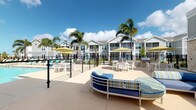 This outdoor amenity space is designed with a resort-inspired aesthetic, centered around a shimmering swimming pool framed by swaying palm trees and a bright Florida sky. The expansive pool deck is finished in a warm, neutral-toned surface that complements the clean, coastal architectural style of the surrounding buildings, which feature gray board-and-batten siding, white trim, and classic gabled rooflines.

The lounge area is furnished with comfortable blue and striped outdoor sofas anchored by natural stone columns, providing a relaxed gathering space just steps from the water. White patio tables and chairs are arranged throughout the deck, ideal for casual outdoor dining and socializing. Umbrella-shaded seating clusters offer additional spots to unwind under the sun.

A clubhouse building anchors one side of the deck, featuring a large multi-pane glass garage-style door that opens to reveal an interior dining and lounge space, seamlessly connecting indoor and outdoor living. A water fountain station and clearly posted pool signage reflect thoughtful amenity planning. Black iron fencing encloses the pool area, adding a polished, structured look. The entire space captures a vibrant, sun-soaked atmosphere well-suited for community gatherings and leisure.