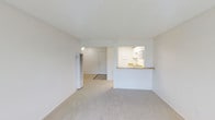 This single-floor unit presents an open and functional layout designed for comfortable everyday living. The main living area is finished with wall-to-wall carpeting in a neutral tone, contributing to a clean and understated aesthetic throughout the space. White walls and a textured ceiling enhance the sense of brightness, keeping the interior feeling airy and open.

A pass-through kitchen cutout connects the kitchen to the living area, promoting an open feel between the two spaces. The kitchen is equipped with under-cabinet lighting, white cabinetry, and a countertop with a warm-toned finish, providing a practical and well-organized cooking space. A built-in microwave is visible above the counter, adding to the kitchen's functionality.

Adjacent to the main living area, a hallway leads to the unit's entry door, which is illuminated by overhead lighting. A wall-mounted heater is positioned along one wall of the living area, providing a dedicated heating source. The overall ambiance of the space is simple, clean, and move-in ready, suited for those seeking a straightforward and low-maintenance living environment.