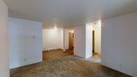 This single-floor space is characterized by a clean, straightforward layout with clearly defined zones. The main living area features neutral-toned carpet, providing a comfortable underfoot surface, while an adjoining room transitions to hard surface flooring, adding functional variety. White walls throughout create a bright, open atmosphere, complemented by a textured ceiling finish. Flush-mounted ceiling light fixtures provide consistent illumination across the space.

A structural column near the center of the space helps define the transition between the carpeted and hard-floored areas without fully separating them, maintaining an open feel. Baseboard heating runs along the wall in the hard-floored section, indicating a reliable, built-in heating system. In the background, warm-toned wooden closet doors are visible, offering dedicated storage space. A second opening reveals an additional area with its own closet, suggesting potential use as a bedroom or flexible-use room. The overall design is practical and understated, with a neutral color palette that provides a versatile backdrop suitable for a variety of furnishing styles.