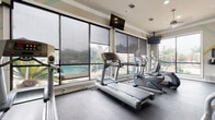 This fitness center is a thoughtfully designed, single-floor workout facility that offers a comprehensive range of exercise equipment in a bright, open environment. Large floor-to-ceiling windows with adjustable roller shades line the perimeter, flooding the space with natural light while providing scenic views of an outdoor pool and lush palm tree landscaping. The cardio zone features multiple treadmills and elliptical machines, each positioned to take full advantage of the poolside views, while wall-mounted flat-screen televisions offer additional entertainment during workouts. The strength training area is equally well-appointed, housing a full rack of dumbbells, a multi-station cable machine, a leg press, an adjustable weight bench, and a power rack, catering to a wide variety of training styles and fitness levels. Full-length mirrors span entire walls throughout the space, visually expanding the room and allowing users to monitor their form. Colorful accent details on the upper walls add a lively, energetic touch to the otherwise neutral palette. The floors are covered in durable, commercial-grade rubber flooring, providing safety and comfort underfoot. Recessed lighting and ceiling fans round out the functional, well-maintained ambiance of this inviting fitness space.