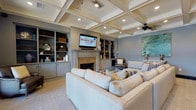 This elegantly designed single-floor lounge space immediately impresses with its distinctive coffered ceiling, which adds architectural depth and a classic, refined character to the room. Recessed lighting and a ceiling fan are seamlessly integrated into the ceiling grid, ensuring the space is both well-lit and comfortable year-round.

The room is anchored by a stone-surround fireplace, above which a wall-mounted flat-screen television is centrally positioned for optimal viewing. Flanking the fireplace on both sides are custom built-in shelving units and lower cabinetry, painted in a soft blue-grey tone that complements the muted wall color throughout. The shelves are tastefully styled with decorative objects, baskets, and books, contributing to a curated yet approachable aesthetic.

Seating is generous and inviting, centered around a large neutral-toned sectional sofa accented with patterned throw pillows. Additional seating includes leather accent chairs and vibrant yellow wingback chairs that introduce a bold pop of color. A glass-top coffee table grounds the central seating arrangement.

A Greek key-patterned area rug defines the main gathering zone, adding visual interest underfoot. French doors on one end of the room allow natural light to filter in and provide access to an outdoor area. An adjacent wet bar area is visible through an open doorway, enhancing the space's entertaining capabilities.
