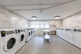 Located at 100 Cosburn Ave in Toronto's East York neighbourhood, this shared laundry facility is a well-organized and efficiently designed space. The room is finished with large-format, light-toned tile on the walls, lending a clean and modern aesthetic throughout. The floor features a distinctive herringbone-patterned tile that adds a refined visual detail underfoot.

The space is equipped with multiple front-loading washing machines and top-loading dryers, arranged along two opposing walls to maximize usable floor space. A stainless steel utility sink is also present, offering additional functionality. At the centre of the room, a stainless steel folding table with a lower shelf provides a practical surface for sorting and folding laundry.

Natural light enters through a row of windows fitted with roller blinds along one wall, creating a bright and pleasant environment. The space is further illuminated by modern circular flush-mount ceiling fixtures that provide even, consistent lighting. A bulletin board near the windows displays relevant building notices and laundry guidelines.

Ventilation is supported by wall-mounted grilles, and the overall layout ensures ease of movement and accessibility throughout. The space reflects a thoughtfully maintained and functional shared amenity, suited to the needs of a residential building.