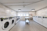 Located at 100 Cosburn Ave in Toronto's East York neighbourhood, this shared laundry facility is a well-organized and efficiently designed space. The room is finished with large-format, light-toned tile on the walls, lending a clean and modern aesthetic throughout. The floor features a distinctive herringbone-patterned tile that adds a refined visual detail underfoot.

The space is equipped with multiple front-loading washing machines and top-loading dryers, arranged along two opposing walls to maximize usable floor space. A stainless steel utility sink is also present, offering additional functionality. At the centre of the room, a stainless steel folding table with a lower shelf provides a practical surface for sorting and folding laundry.

Natural light enters through a row of windows fitted with roller blinds along one wall, creating a bright and pleasant environment. The space is further illuminated by modern circular flush-mount ceiling fixtures that provide even, consistent lighting. A bulletin board near the windows displays relevant building notices and laundry guidelines.

Ventilation is supported by wall-mounted grilles, and the overall layout ensures ease of movement and accessibility throughout. The space reflects a thoughtfully maintained and functional shared amenity, suited to the needs of a residential building.