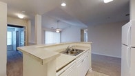 This single-floor residence showcases a bright, open-concept design that seamlessly connects the kitchen and living areas. The kitchen is anchored by a generously sized peninsula with light-toned countertops, white cabinetry, and a double-basin stainless steel sink — offering both ample prep space and a convenient breakfast bar. Recessed lighting above keeps the kitchen well-illuminated, complementing the natural light that filters in through large covered windows in the adjoining living area. The living space features plush wall-to-wall carpeting and is crowned by soaring vaulted ceilings that lend an airy, expansive feel to the interior. A ceiling fan with a light fixture hangs centrally, providing both comfort and ambient lighting. Structural pillars subtly define the transition between spaces while maintaining the open flow of the floor plan. A hallway visible from the main living area leads to additional rooms, suggesting a thoughtful layout designed for comfortable everyday living. The neutral color palette throughout — soft whites, creams, and warm beiges — creates a clean, versatile backdrop well-suited to a variety of interior styles. Tile flooring in the kitchen transitions smoothly to carpet in the living zone, adding practical definition between the two areas.