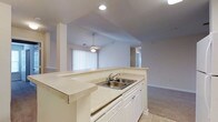 This single-floor residence showcases a bright, open-concept design that seamlessly connects the kitchen and living areas. The kitchen is anchored by a generously sized peninsula with light-toned countertops, white cabinetry, and a double-basin stainless steel sink — offering both ample prep space and a convenient breakfast bar. Recessed lighting above keeps the kitchen well-illuminated, complementing the natural light that filters in through large covered windows in the adjoining living area. The living space features plush wall-to-wall carpeting and is crowned by soaring vaulted ceilings that lend an airy, expansive feel to the interior. A ceiling fan with a light fixture hangs centrally, providing both comfort and ambient lighting. Structural pillars subtly define the transition between spaces while maintaining the open flow of the floor plan. A hallway visible from the main living area leads to additional rooms, suggesting a thoughtful layout designed for comfortable everyday living. The neutral color palette throughout — soft whites, creams, and warm beiges — creates a clean, versatile backdrop well-suited to a variety of interior styles. Tile flooring in the kitchen transitions smoothly to carpet in the living zone, adding practical definition between the two areas.