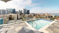 Located at 550 W Date St in the heart of downtown San Diego, this rooftop amenity deck offers a compelling outdoor retreat with panoramic views of the city skyline, San Diego Bay, and surrounding neighborhoods. The expansive tiled terrace is thoughtfully divided into distinct zones, each designed for a different aspect of outdoor living.

A sparkling rooftop pool anchors one section of the deck, framed by glass railings that preserve the breathtaking views of downtown towers and the bay beyond. Adjacent to the pool, sun loungers with bold red cushions invite relaxation beneath the open California sky, while large planters with manicured olive trees add a touch of greenery and privacy.

The social hub of the rooftop features a full-length bar lined with contemporary high-top seating, ideal for gatherings. Nearby, a covered lounge area with plush sectional sofas and a central fire pit creates a warm, intimate setting for evening conversations. Overhead, string lights and large shade canopies provide comfort and ambiance day and night.

An outdoor kitchen equipped with built-in appliances, a grill, and ample counter space supports al fresco dining and entertaining. Glass railings throughout the deck maintain unobstructed sightlines to the San Diego skyline, making this rooftop a premier destination for outdoor amenity and city living.