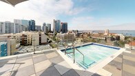 Located at 550 W Date St in the heart of downtown San Diego, this rooftop amenity deck offers a compelling outdoor retreat with panoramic views of the city skyline, San Diego Bay, and surrounding neighborhoods. The expansive tiled terrace is thoughtfully divided into distinct zones, each designed for a different aspect of outdoor living.

A sparkling rooftop pool anchors one section of the deck, framed by glass railings that preserve the breathtaking views of downtown towers and the bay beyond. Adjacent to the pool, sun loungers with bold red cushions invite relaxation beneath the open California sky, while large planters with manicured olive trees add a touch of greenery and privacy.

The social hub of the rooftop features a full-length bar lined with contemporary high-top seating, ideal for gatherings. Nearby, a covered lounge area with plush sectional sofas and a central fire pit creates a warm, intimate setting for evening conversations. Overhead, string lights and large shade canopies provide comfort and ambiance day and night.

An outdoor kitchen equipped with built-in appliances, a grill, and ample counter space supports al fresco dining and entertaining. Glass railings throughout the deck maintain unobstructed sightlines to the San Diego skyline, making this rooftop a premier destination for outdoor amenity and city living.