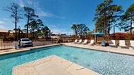 Located in Gulf Shores, Alabama, this single-story pool facility offers a well-appointed outdoor and semi-enclosed environment designed for relaxed coastal living. The centerpiece is a sparkling swimming pool with light blue water, framed by a stamped concrete surround and a classic wrought iron safety fence. The pool deck is furnished with white lounge chairs and a teal umbrella-shaded seating area, providing ample space for sunbathing and socializing beneath the open Alabama sky. Mature pine and palm trees line the perimeter, adding natural character to the setting.

Adjacent to the pool, a screened enclosure serves as a comfortable gathering space. Large floor-to-ceiling screen windows wrap the room, flooding the interior with natural light while offering unobstructed views of the pool and surrounding landscape. The space features coffered ceilings with recessed lighting and clean crown molding, lending a polished, finished feel. Horizontal shiplap-style siding in a soft sage-gray hue lines the interior walls, complementing the bright, airy atmosphere.

A dedicated wet bar area with dark cabinetry, a sink, and granite-style countertops adds functional convenience. A round metal dining table with chairs provides an additional seating option. Multiple entry doors connect the interior room directly to the outdoor pool deck, creating seamless indoor-outdoor flow ideal for gatherings.