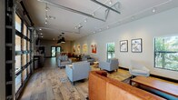 This single-story space is designed as a contemporary communal gathering area, blending industrial and modern design elements across an open, elongated floor plan. Wide-plank hardwood flooring flows throughout, unifying the distinct zones within the space. High ceilings with exposed structural elements and track lighting provide ample illumination, while large windows along the perimeter invite natural light and offer views of lush tropical greenery outside.

The main lounge area features multiple seating arrangements, including modular gray sofas and armchairs grouped atop patterned area rugs, creating intimate conversation zones. A standout textured brick accent wall anchors one end of the lounge, paired with a wall-mounted flat-screen television and complemented by curated abstract artwork displayed along the white walls.

A dedicated kitchenette area is outfitted with dark cabinetry, white countertops, a stainless steel refrigerator, a sink, and a wall of white subway tile. A mounted TV and casual high-top dining table with industrial-style seating make this area equally functional for dining or socializing. Beyond the kitchenette, a workstation nook with bar-height seating provides a practical area for focused tasks. The overall ambiance is welcoming, modern, and well-appointed for community gathering and collaboration.