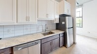 Located at 104 E Second St in Richmond, Virginia, this single-floor unit masterfully blends industrial character with modern finishes. The space is defined by striking exposed brick walls that lend a warm, textured aesthetic throughout, complemented by polished concrete floors that reinforce the loft-inspired design language.

The kitchen is well-appointed with a full suite of stainless steel appliances, including a Frigidaire refrigerator, dishwasher, microwave, and range. White shaker-style upper cabinets contrast elegantly with dark lower cabinetry, while light granite countertops and a classic white subway tile backsplash complete the contemporary look. Exposed ductwork overhead adds to the industrial ambiance of the kitchen area.

The bathroom features a clean, modern design with full-height white tile surrounding a soaking tub and shower combo. Brushed nickel fixtures, including a curved shower rod and integrated hardware, provide a polished finishing touch. A secondary bathroom mirrors this layout with the same crisp tiling and brushed nickel detailing.

The open living areas showcase the raw architectural character of the building, with soaring exposed brick walls and generous ceiling height that create a sense of volume and openness. This space offers a compelling combination of historic industrial charm and thoughtfully updated modern amenities.