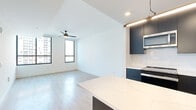This contemporary 2-bedroom, 1-bathroom unit is thoughtfully designed with a clean, modern aesthetic throughout. Light hardwood-style flooring flows seamlessly across the entire space, complementing the crisp white walls and creating a bright, cohesive feel.

The open-concept kitchen is a standout feature, showcasing sleek dark cabinetry that extends to the ceiling, a white quartz waterfall island, white subway tile backsplash, and a full suite of stainless steel appliances including a refrigerator with ice maker, range, dishwasher, and microwave. Pendant lighting above the island adds a refined touch.

The kitchen opens directly into the living area, which benefits from natural light through large black-framed windows and is equipped with a ceiling fan. Both bedrooms continue the modern theme with light hardwood floors, ceiling fans, and expansive black-framed windows that frame urban cityscape views. The primary bedroom features tall windows on two walls, filling the room with abundant natural light.

The bathroom is finished to a high standard, featuring a glass-enclosed walk-in shower with white subway tile, an LED-backlit vanity mirror, dark cabinetry with a white countertop, and wood-look flooring. Two closets provide practical storage, and a hallway connects the living spaces efficiently.