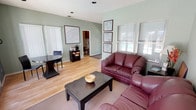 This single-floor community or leasing office is designed to be both functional and inviting, offering a well-organized layout that accommodates visitors with ease. The interior is finished with warm light wood flooring throughout the main areas, complemented by soft sage green walls that create a calm, professional atmosphere. A textured ceiling with recessed lighting provides consistent illumination across the space.

The main lounge area is furnished with deep burgundy leather loveseats arranged around a dark wood coffee table, providing a comfortable setting for guests. Framed floor plan displays are mounted along the walls, offering informative references for prospective residents. A small side table with a lamp and decorative accents adds a welcoming residential touch.

Adjacent to the lounge, a compact refreshment station with a coffee maker and snack display serves visitors. A wide hallway with a glass-paneled entry door leads into the space, featuring a reception counter area with framed community maps and plan layouts prominently displayed.

A standout feature of the space is the back wall, finished in a rich dark teal, anchored by two sets of white-framed French doors that open directly to an outdoor pool area, flooding the interior with natural light and offering clear views of the exterior amenity space. A small dining-style table and chairs near the French doors provide an additional functional area within this bright, open setting.