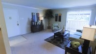 This single-floor residence offers a comfortable and well-appointed living environment designed with both style and function in mind. The living room serves as the heart of the space, featuring soft wall-to-wall carpeting that adds warmth underfoot and a clean, neutral color palette that creates a calm and inviting atmosphere. Crown molding lines the ceiling, lending a refined architectural touch to the room.

Natural light pours in through a large window dressed with vertical blinds, offering a view of the outdoors and brightening the interior throughout the day. The living area is generously sized, easily accommodating a full furniture arrangement that includes a central coffee table, an accent chair, and a sideboard with decorative accessories. A tall indoor plant in the corner adds a natural, organic element to the space.

Wall art in bold, contrasting tones provides visual interest and personality to the otherwise understated walls. The overall ambiance is relaxed yet polished, striking a balance between everyday livability and thoughtful interior design. The single-level layout ensures easy accessibility and a seamless flow throughout, making this a practical and appealing space for comfortable day-to-day living.