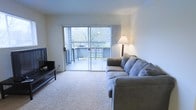This single-floor residence offers a warm and welcoming layout designed for comfortable everyday living. The living room serves as the heart of the space, featuring plush wall-to-wall carpeting that adds a cozy feel underfoot. A large sliding glass door floods the room with natural light and provides direct access to a private outdoor deck, where views of surrounding trees and lush greenery create a serene, nature-connected atmosphere. Sheer vertical blinds frame the glass door, allowing for flexible light control while maintaining the open, airy feel of the room. The living area comfortably accommodates a generous sectional sofa and an entertainment setup, making it well-suited for both relaxing and hosting. A floor lamp provides warm ambient lighting, complementing the natural light that fills the space throughout the day. The neutral-toned walls and light carpeting create a clean, versatile backdrop that adapts easily to a variety of décor styles. Overall, the space strikes a balance between functionality and comfort, with its indoor-outdoor connection being a notable highlight that enhances the overall ambiance.