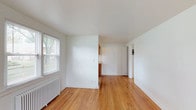 Unit 14 at Warner Village Apartments is a single-story residence that presents a clean, classic aesthetic throughout. Warm-toned hardwood floors run continuously across the living areas and bedrooms, lending a timeless character to the space. Crisp white walls and white-trimmed baseboards create a bright, neutral backdrop throughout.

The living area is well-proportioned, featuring multiple windows that invite natural light and views of the surrounding greenery. A cast-iron radiator provides traditional heating, adding a classic architectural detail. The space opens toward a dedicated kitchen area, maintaining an accessible flow between rooms.

The kitchen is efficiently laid out in a galley configuration, featuring warm wood cabinetry, a tile backsplash, light countertops, a stainless steel sink, and a window above the radiator that brings in additional natural light. White appliances, including a gas range and full-sized refrigerator, are included.

The bedroom is generously sized, featuring dual windows, hardwood flooring, and a radiator, with access to a closet and the broader hallway. The bathroom is finished with neutral tile surrounds, a combination tub and shower, and a window for ventilation. Overall, the unit offers a straightforward, well-maintained living environment with consistent finishes throughout.