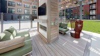 This thoughtfully designed outdoor amenity space serves as a vibrant communal hub set within a modern residential development. The centerpiece of the space is a covered deck area framed by a wood-clad pergola structure, offering a semi-sheltered lounge environment. A built-in fireplace with a wall-mounted television anchors the deck, creating an inviting focal point for gathering. Contemporary outdoor seating — including green cushioned sofas and distinctive red woven accent chairs — adds both comfort and visual interest to the space.

The deck transitions to an expansive open-air lawn, lush with well-maintained green grass that provides a generous open area for recreation and relaxation. Concrete pathways border the lawn, connecting the various zones of the courtyard seamlessly. Raised planter beds with ornamental shrubs and young trees introduce natural greenery and softscape throughout, complementing the surrounding modern architecture.

The courtyard is enclosed by multi-story residential buildings clad in a mix of red brick, gray panel siding, and light-toned facades, creating a sense of enclosure and community. String lights hint at evening ambiance, while the open sky above ensures abundant natural light throughout the day. The overall design balances functionality with aesthetic appeal, offering a welcoming outdoor retreat.