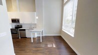 This single-floor space showcases a clean, contemporary design that emphasizes openness and functionality. The kitchen is outfitted with crisp white cabinetry, stone-look countertops, and stainless steel appliances, creating a polished and efficient cooking area. A pendant light fixture hangs above the extended countertop, which doubles as a casual dining or workspace surface, adding both style and versatility to the layout.

The adjoining dining area benefits from a large window dressed with horizontal blinds, allowing generous natural light to filter in and illuminate the rich, dark hardwood-style flooring that runs throughout the space. The warm tones of the flooring provide a striking contrast against the neutral, light-toned walls and white trim, contributing to a balanced and inviting ambiance.

The ceilings are notably tall, enhancing the sense of volume and airiness throughout the open-plan area. The neutral color palette — anchored by soft beige walls and bright white accents — creates a versatile backdrop suitable for a variety of interior styles. Overall, the space combines modern finishes with thoughtful design, offering a bright, well-appointed environment that feels both comfortable and sophisticated.