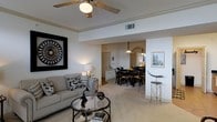 This single-floor residence offers an inviting and cohesive living environment defined by a seamless open-plan layout that connects the main living area to the dining space. The living room is furnished with a classic neutral-toned sofa accented with patterned throw pillows, centered around a glass-top coffee table. A ceiling fan with a light fixture overhead provides both comfort and illumination, while bold wall art adds a striking visual focal point. Tray ceiling detailing lends architectural interest and a sense of refined height to the room. Light-toned carpeting runs throughout, contributing to the warm and approachable ambiance. The dining area, visible through an open passageway, features a dark wood dining table surrounded by matching chairs, complemented by a decorative pendant light above. The kitchen is partially visible beyond the dining space, hinting at a practical and connected layout. Wall-mounted artwork and decorative accents are thoughtfully placed throughout, reinforcing a polished, curated aesthetic. Natural and artificial lighting work in harmony to create a bright, welcoming atmosphere across the entire living space.