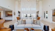 This single-floor space makes a striking architectural impression through its dramatic double-height ceilings and an open, airy layout that flows seamlessly between distinct functional zones. The main lounge area is anchored by a towering floor-to-ceiling marble slab wall housing a sleek linear fireplace, creating a bold focal point. Neutral gray sofas, cognac leather wingback chairs, and geometric accent pillows furnish the lounge with refined contemporary style, while light wood-look tile flooring runs throughout, unifying the space.

A dedicated bar and gathering area features a generously sized white quartz island with seating for several guests, backed by a distinctive geometric wood-and-dark-tile accent wall. Clerestory windows and glass-paned doors flood this area with abundant natural light. An adjacent entry zone showcases large floor-to-ceiling windows overlooking greenery, framing a decorative console table beneath a bold multi-panel artwork.

The office area offers a purposeful workspace with two dark desks, modern wood-and-metal guest chairs, and warm sputnik-style pendant lighting, complemented by patterned carpet tile flooring. Throughout the entire space, the design balances elegance with functionality, creating a welcoming, upscale environment suited for community gathering and professional interaction.